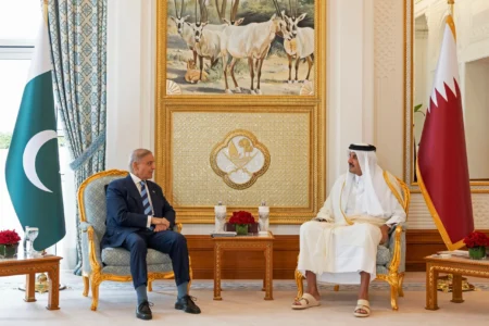 An official indoor meeting showing Pakistan Prime Minister Shehbaz Sharif in a dark business suit seated in an ornate armchair conversing with Qatar Emir Sheikh Tamim bin Hamad Al Thani in traditional Qatari attire, flanked by the national flag of Pakistan on the left and the flag of Qatar on the right, highlighting regional mediation efforts tied to Iran-U.S. Diplomacy.