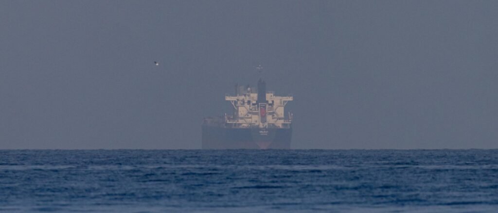 What a Fragile U.S.-Iran Ceasefire Means for Energy and Beyond A distant view of a large cargo ship or oil tanker navigating across a wide, hazy blue sea horizon with a single bird flying in the clear sky, representing energy trade corridors monitored during a U.S.-Iran Ceasefire.