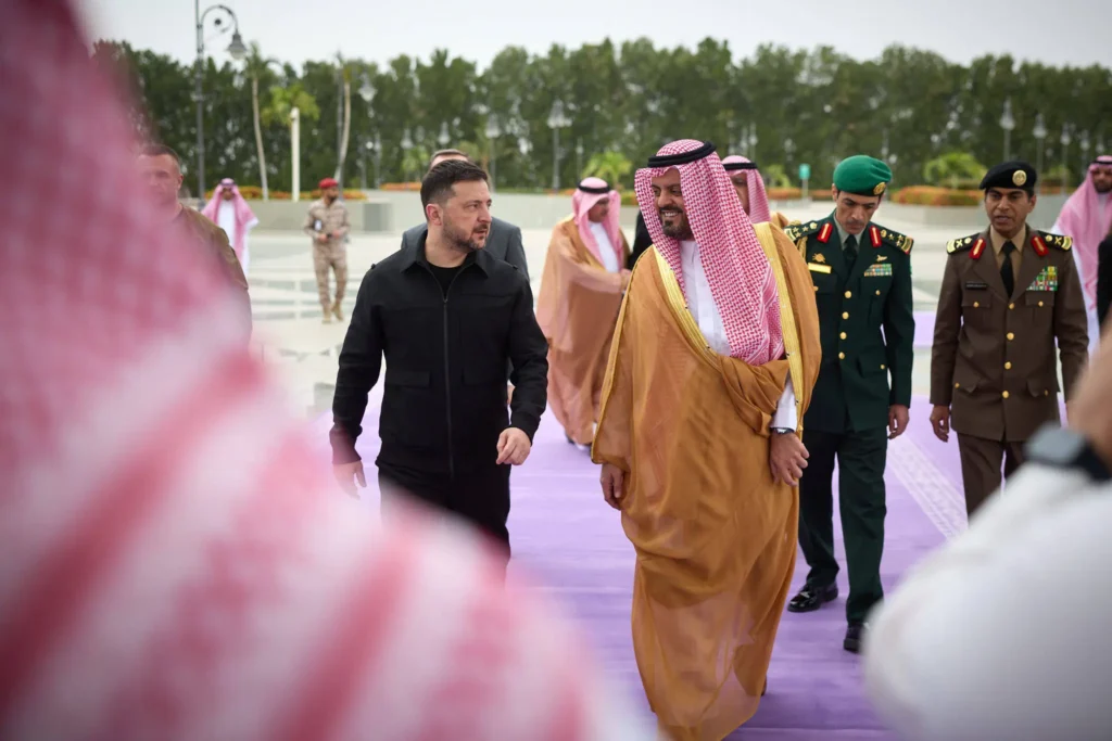 Ukrainian President Volodymyr Zelenskyy walks along a purple carpet alongside a smiling Saudi official in traditional attire, accompanied by military officers and delegates outdoors, illustrating diplomatic partnerships between Europe and the Gulf.