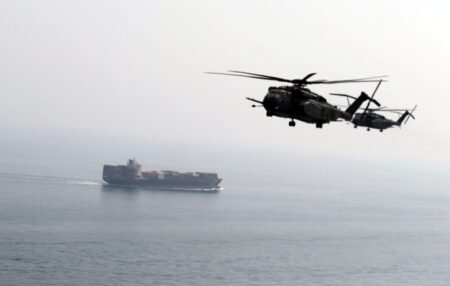 A wide-angle view of two military helicopters flying in formation over the sea, with a large container cargo ship sailing in the background under a hazy sky, illustrating maritime security exercises and naval operations linked to Sea Mines.