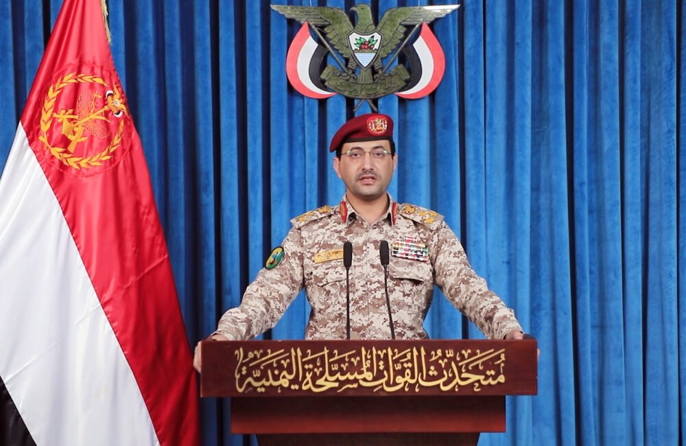 Iran’s Axis of Resistance Strategy Targets Key Chokepoints A Houthi military spokesperson in camouflage fatigues delivering a press statement behind a podium beside the Yemeni flag, representing regional military factions tied to the Iran axis of resistance.