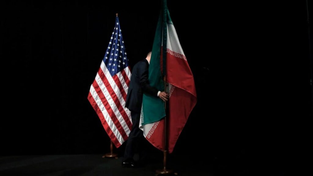 Why Hasn’t the Trump Administration Complied with the Law on Reporting Iran Agreements to Congress? A man in a dark suit walks behind a large national flag of Iran positioned next to a United States flag against a stark black background, symbolizing clandestine diplomatic maneuvers related to Iran agreements.