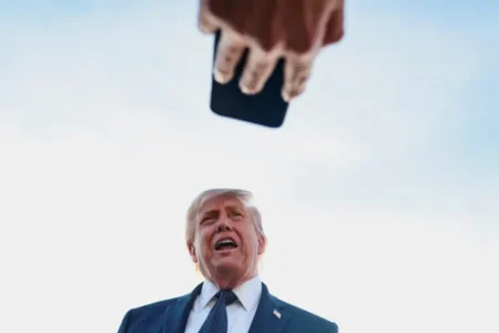 A low-angle, close-up shot of U.S. President Donald Trump speaking to the media, with a hand holding a smartphone positioned in the foreground, visually representing the modern challenges and media dynamics surrounding Trump's Foreign Policy.