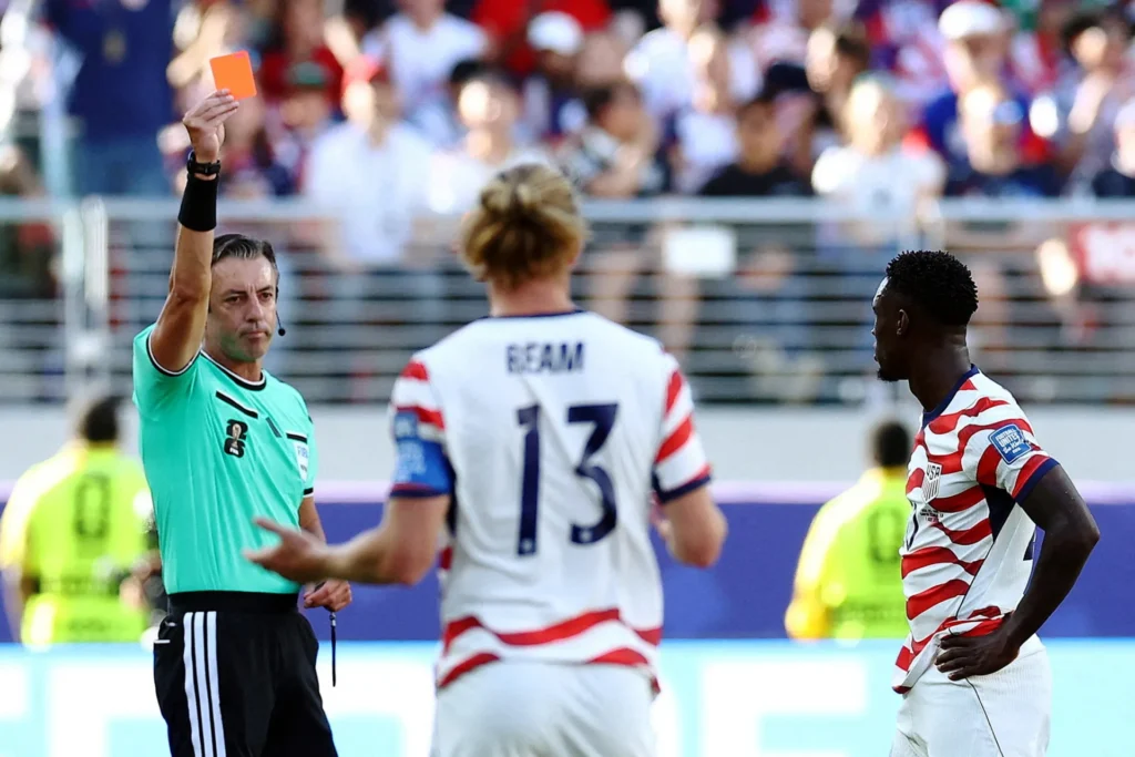 A referee in a light green uniform holds up a red card in front of U.S. Men's National Team player Folarin Balogun and teammate Tim Ream during a soccer match with a blurred crowd in the background, illustrating the controversy surrounding the Balogun Red Card Reversal.