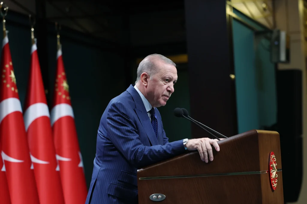 كيف يمنح ترامب وقمة الناتو الشرعية للرئيس التركي الاستبدادي Turkish President Recep Tayyip Erdogan in a pinstripe suit leans forward at a wooden podium speaking into microphones, with multiple national flags of Turkey in the background, framing international relations involving Turkey's Autocratic President.