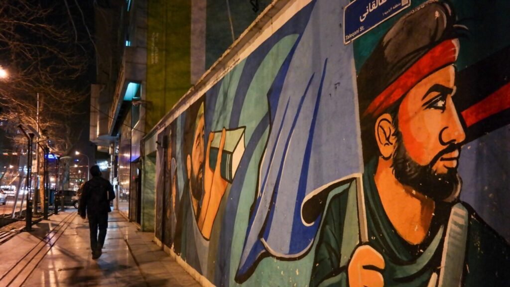 A lone pedestrian walks down a city sidewalk at night next to a long concrete wall covered in hand-painted pro-Islamic Republic murals in Tehran, with a street sign for Taleqani St, framing regional ideological analysis regarding Iran's Terrorist Project.