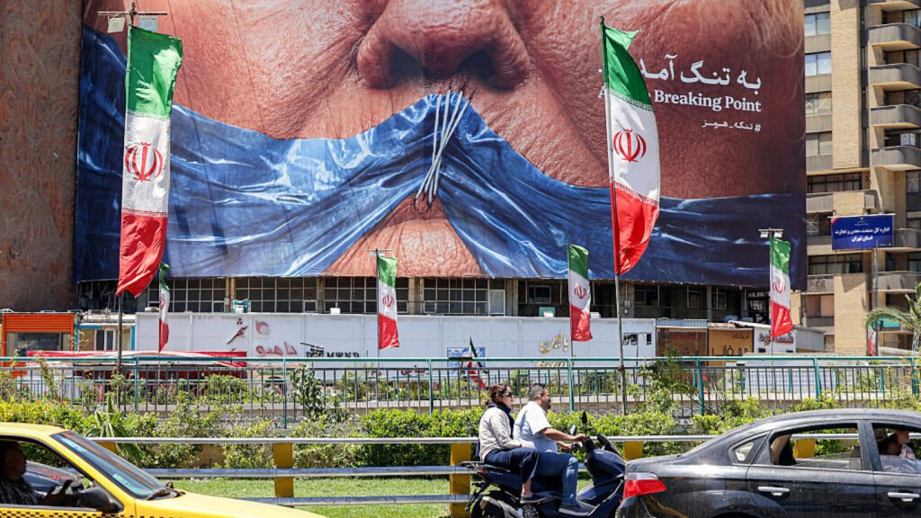 How Trump is trying to rewrite the Iran deal on the battlefield A Tehran street scene shows Iranian flags in front of a large billboard depicting a torn blue mask over a face, symbolizing Iran's political crisis and Trump's efforts to rewrite the Iran deal through military pressure.