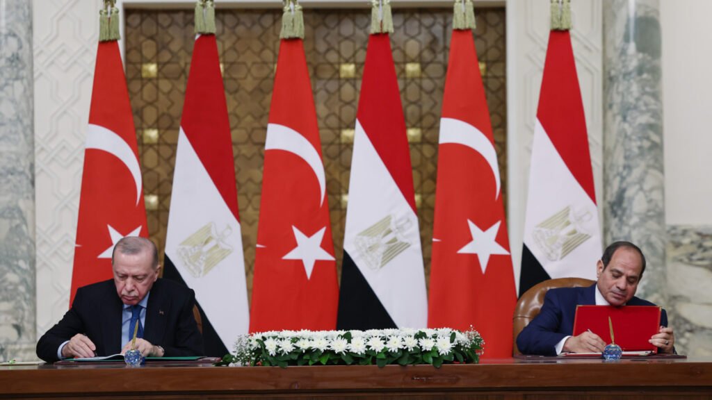 Turkish President Recep Tayyip Erdogan and Egyptian President Abdel Fattah el-Sisi seated at a long wooden table signing official documents, with alternating national flags of Turkey and Egypt lined up in the background, representing bilateral agreements tied to Egypt-Turkey military cooperation.