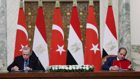Turkish President Recep Tayyip Erdogan and Egyptian President Abdel Fattah el-Sisi seated at a long wooden table signing official documents, with alternating national flags of Turkey and Egypt lined up in the background, representing bilateral agreements tied to Egypt-Turkey military cooperation.