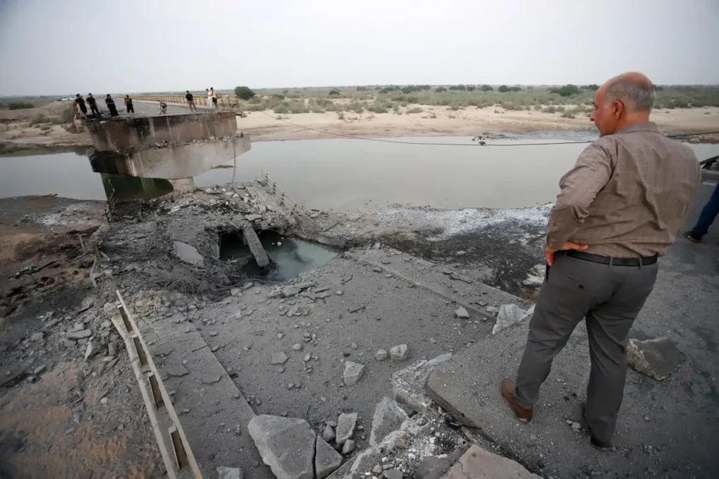 Iran Civilian Infrastructure Strikes Draw War Crimes Scrutiny A man looking over the collapsed concrete section of a destroyed bridge in Hormozgan province, illustrating severe damage to Iran Civilian Infrastructure.