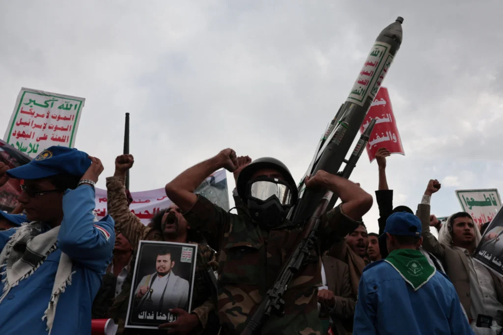 A demonstrator wearing a helmet and goggles holds up a replica missile while surrounded by protesters waving banners at a rally in Sanaa, illustrating tension surrounding the Houthi Blockade.