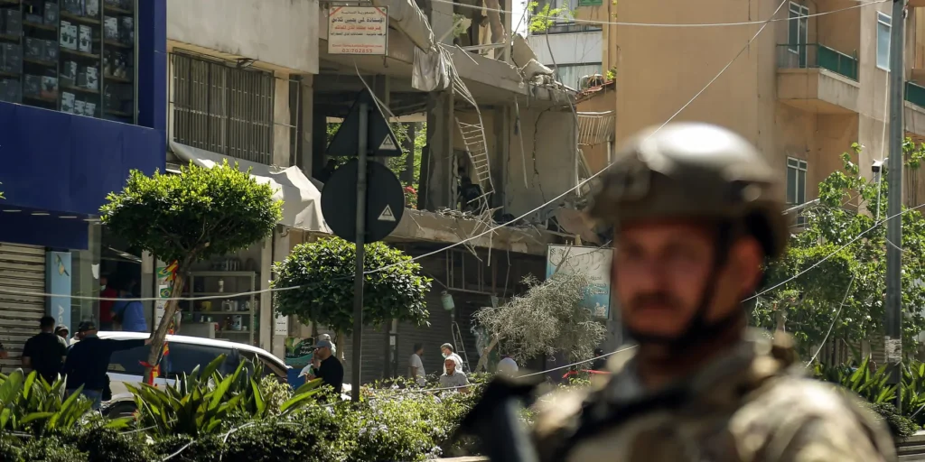 A soldier in tactical gear and a combat helmet stands out of focus in the foreground of a city street where a heavily damaged concrete building with exposed rebar and shattered facades serves as the background, capturing local instabilities and Lebanon Risks.