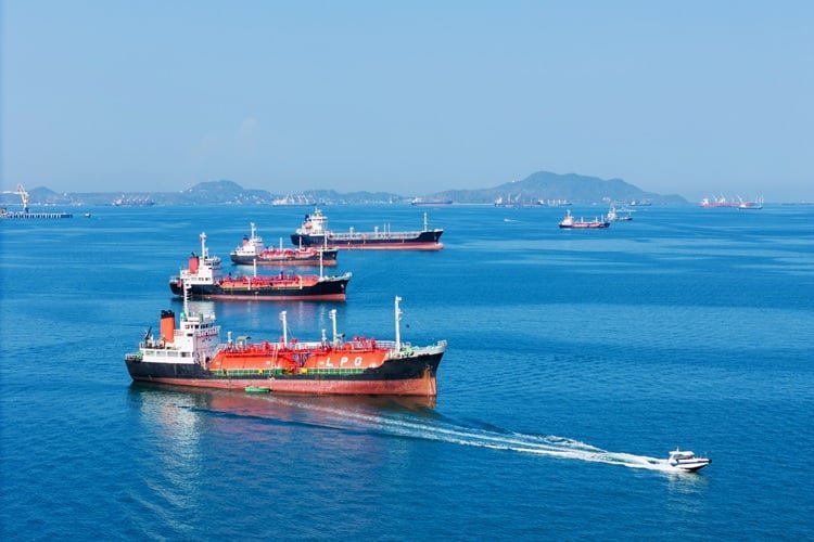 Trump Abandons Freedom of Navigation A high-angle shot of multiple large tankers and commercial ships anchored or sailing on a vibrant blue sea under a clear sky, with a small speed boat cruising in the foreground and hilly coastlines visible in the far background, representing international maritime transit and the Freedom of Navigation.