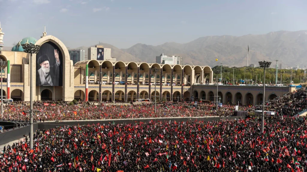 A high-angle view of a massive crowd gathering in the large courtyard of the Imam Khomeini Grand Mosalla in Tehran, with a giant portrait of the late leader hanging on the main archway as thousands of people wave flags during Khamenei's funeral.