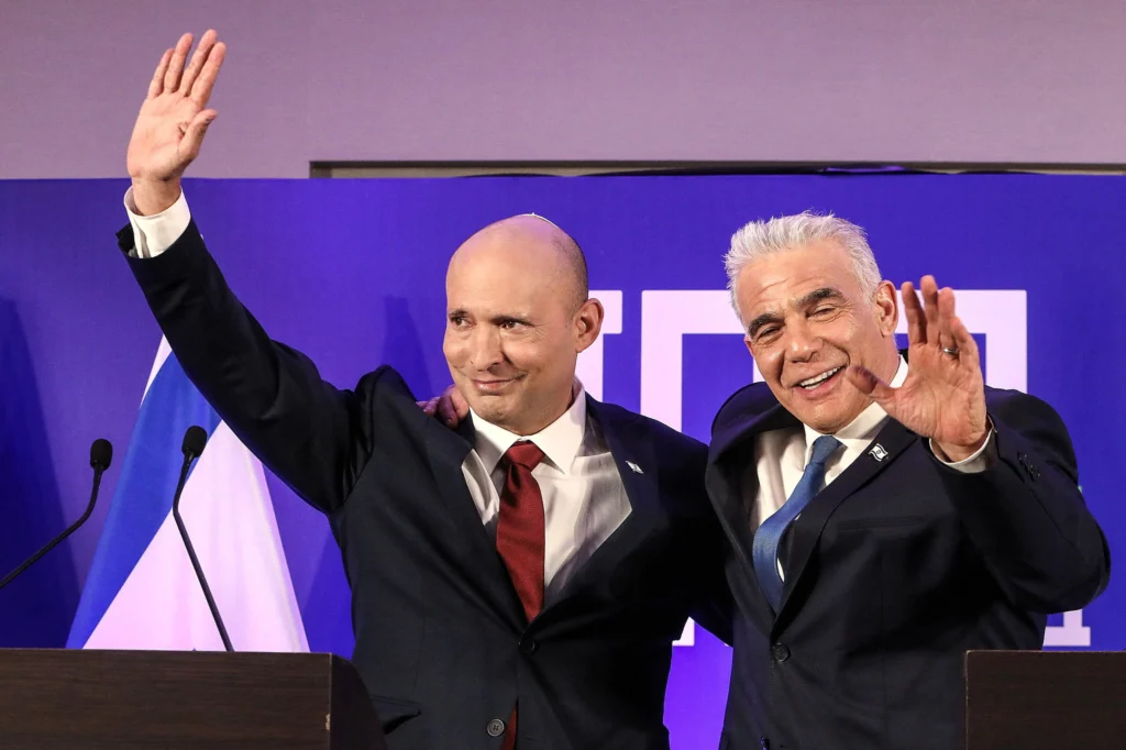 دولت آینده اسرائیل باید به توافقات ابراهیم اولویت دهد A medium shot of Naftali Bennett and Yair Lapid standing side by side in suits at a podium, smiling and waving to the audience with an Israeli flag partially visible in the background, illustrating leadership transitions related to the Abraham Accords.
