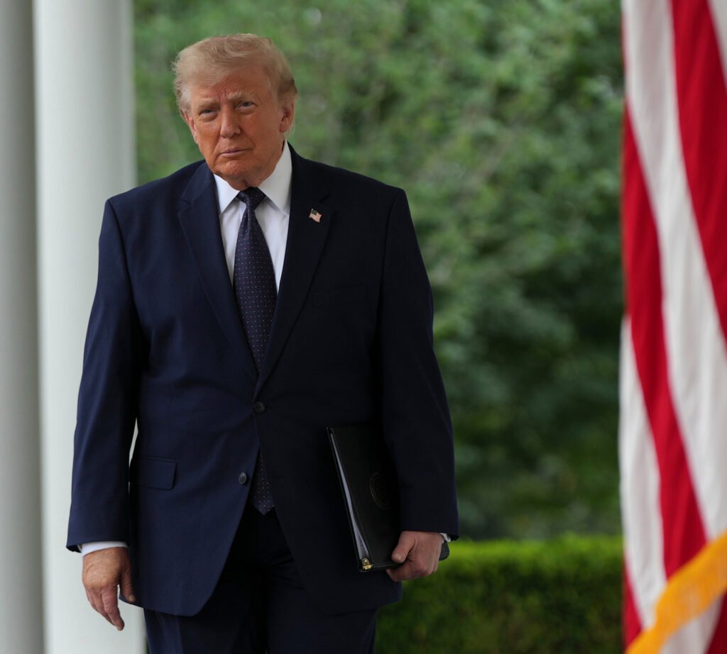 Trump’s Foreign Policy Stumbles After Just 18 Turbulent Months A medium shot of Donald Trump in a navy suit and tie, holding a black folder and walking past a blurred American flag, serving as a visual representation of his administration's Trump's Foreign Policy.