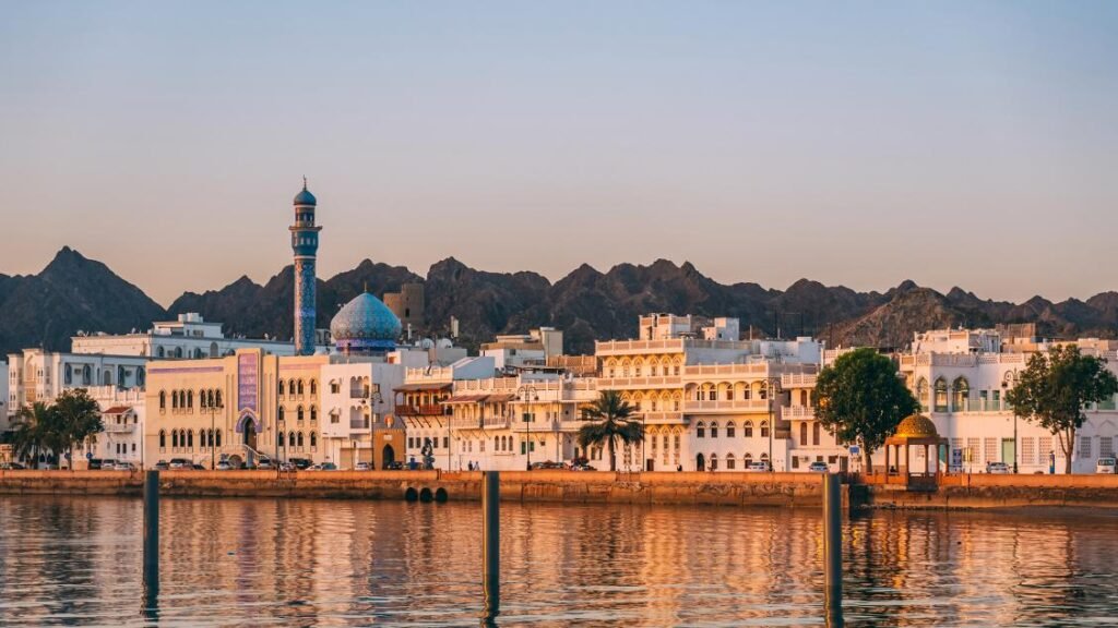 A wide panoramic waterfront view of the Mutrah Corniche in Muscat featuring traditional white buildings, a prominent blue-domed mosque with a tall minaret, and a backdrop of rugged dark mountains reflecting on the calm water at golden hour, illustrating the regional development linked to Sultan Haitham Reforms.