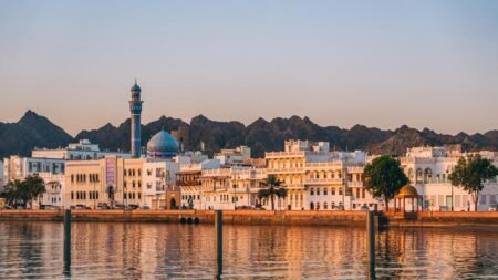 A wide panoramic waterfront view of the Mutrah Corniche in Muscat featuring traditional white buildings, a prominent blue-domed mosque with a tall minaret, and a backdrop of rugged dark mountains reflecting on the calm water at golden hour, illustrating the regional development linked to Sultan Haitham Reforms.