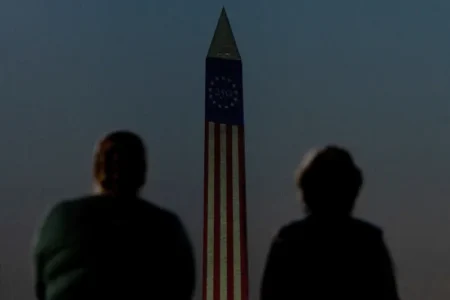 A low-angle shot showing the silhouettes of two people in the foreground looking toward the Washington Monument, which is illuminated with a projection of the American flag stripes and a "250" star circle emblem against a dark twilight sky, capturing symbolic themes connected to America's Global Image.