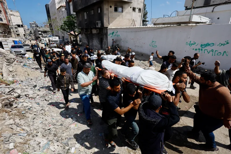 The End of Hamas A large crowd of men carries a body wrapped in a white shroud on an orange stretcher through a rubble-strewn street in Gaza City, depicting the severe toll of conflict and discussions regarding the End of Hamas.