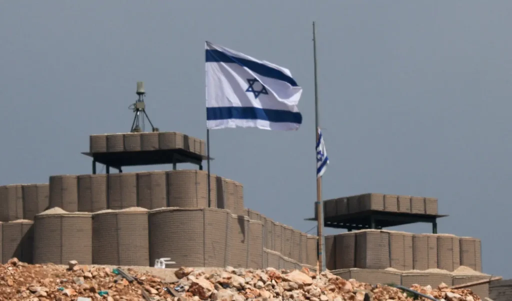 An Israeli flag flutters in the wind at a fortified military base near the border with Lebanon, featuring defensive sandbag barriers and communication equipment against a grey sky, symbolizing the geopolitical tensions surrounding Israel's Isolation.