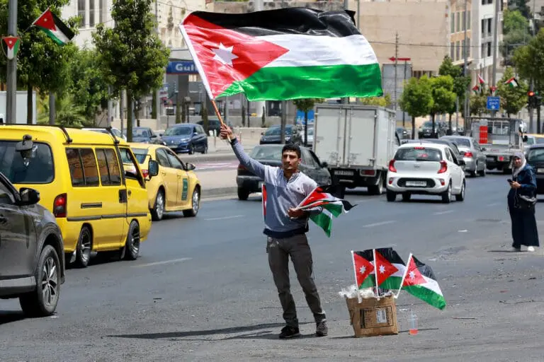 Jordan’s Delicate Path Forward in a Changing Middle East A man stands on the divider of a busy city street with traffic waving a large national flag of Jordan next to a cardboard box filled with smaller flags, capturing the local political backdrop relevant to Jordan's delicate path.