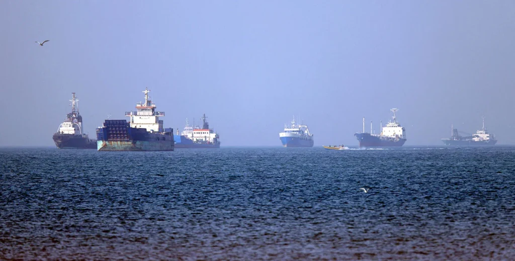 Several commercial cargo ships and maritime vessels are anchored across a wide expanse of choppy blue sea under a hazy sky, highlighting shipping vulnerabilities linked to a regional Gulf Crisis.