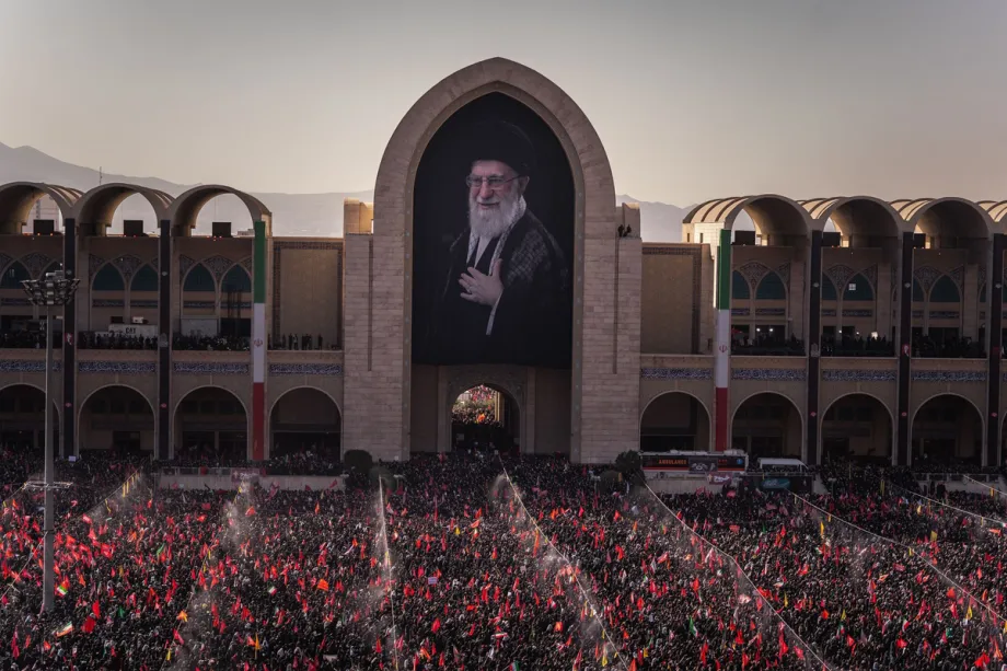 The 4th of July: Tehran’s funeral and Trump’s birthday An overhead view of an immense crowd packed into the courtyard of the Imam Khomeini Mosalla Mosque holding countless flags, beneath a massive archway displaying a portrait of Ali Khamenei, providing the setting for Tehran's funeral.