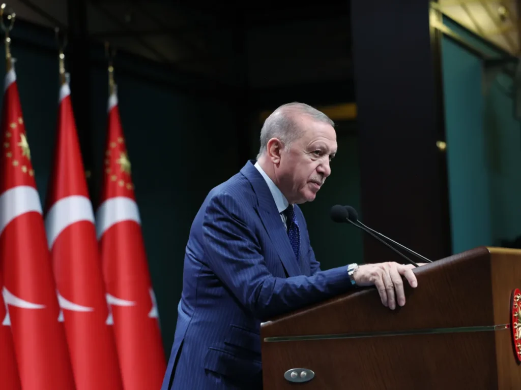 Turkish President Recep Tayyip Erdogan stands in profile at a wooden podium speaking into microphones with Turkish national flags lined up behind him, highlighting the political leadership central to discussions around Erdogan's record.