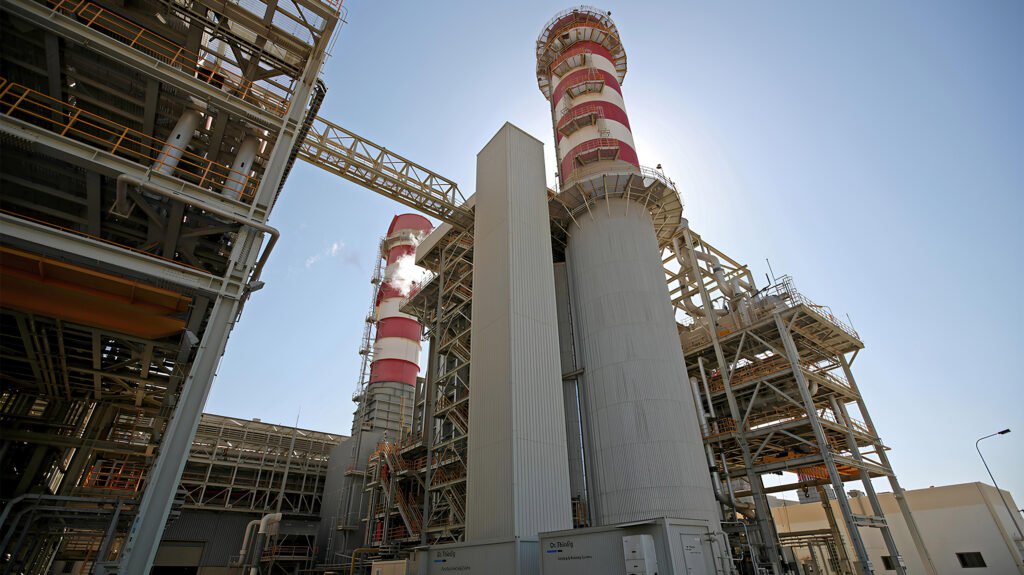 A low-angle view of a large industrial desalination plant featuring tall red-and-white striped exhaust chimneys and complex steel infrastructure, representing critical water facilities tied to Desalination Deterrence.