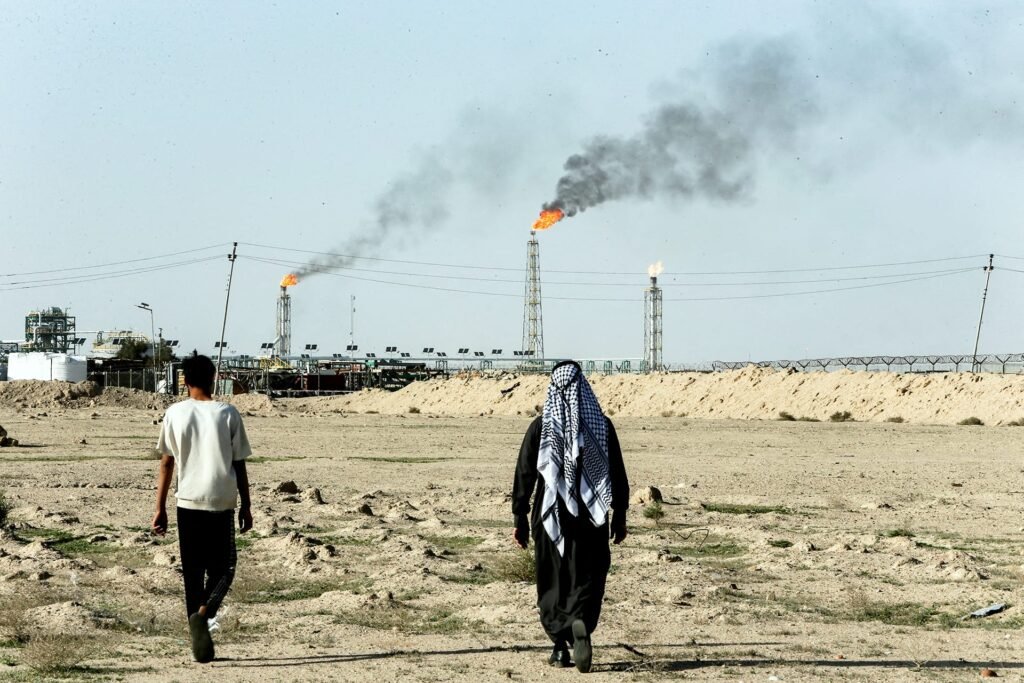 A view from behind of two individuals walking across a dry, barren landscape toward an industrial refinery facility where multiple gas flares burn with smoke in the background, illustrating the regional infrastructure supporting Gulf oil.