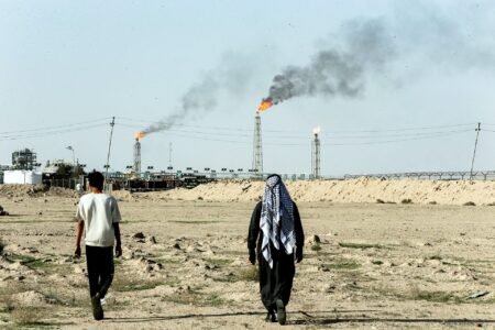 A view from behind of two individuals walking across a dry, barren landscape toward an industrial refinery facility where multiple gas flares burn with smoke in the background, illustrating the regional infrastructure supporting Gulf oil.