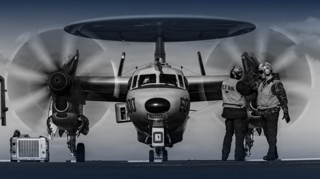 Could the US-Iran war spark World War III? A head-on, black and white shot of a military E-2 Hawkeye airborne early warning aircraft with its propellers spinning on a flight deck as crew members stand nearby, reflecting global military readiness and the geopolitical tensions linked to World War III.