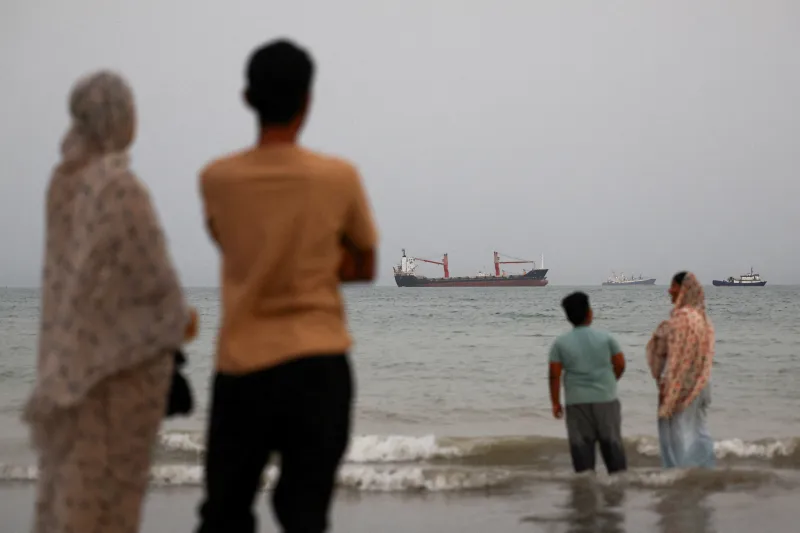 امیدهای امنیتی خلیج-چین پس از جنگ ایران فروپاشید People standing along the shoreline near Bandar Abbas looking out at a cargo ship in the Strait of Hormuz, underscoring maritime concerns tied to Gulf-China Security.