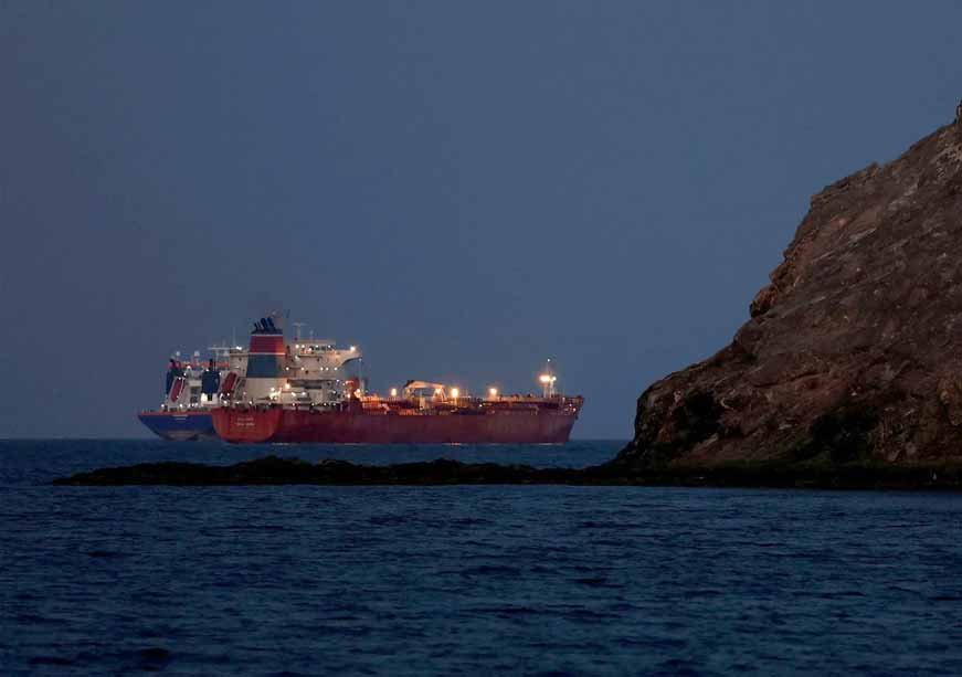 كيف نجت الهند من أكبر صدمة نفطية في التاريخ A side profile view of a large oil tanker vessel with its deck lights turned on navigating through dark blue coastal waters at dusk or night, passing closely behind a rocky cliff formations, symbolizing global energy supply routes during the Biggest Oil Shock.