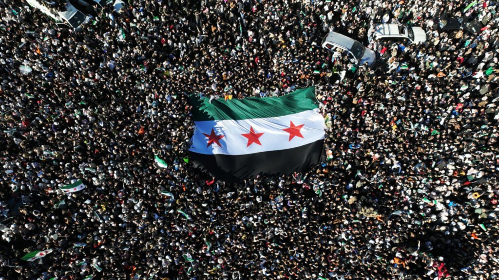 دمشق تحت النار: من يريد فشل السلام الهش في سوريا؟ An aerial top-down view of a massive crowd holding a large Syrian flag with three red stars in the center of a public demonstration, reflecting political stability and Syria's fragile peace.