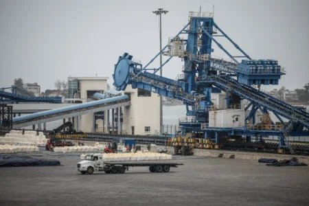 A industrial view of a bustling port terminal featuring a large blue loading crane, conveyor systems, and a transport truck carrying cargo bags, symbolizing the economic infrastructure at the center of the U.S.-China Rivalry.