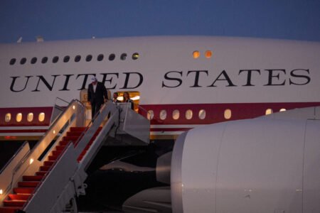 A side profile view of a large white aircraft displaying the prominent text "UNITED STATES" on the fuselage, with individuals descending the mobile airstair passenger steps at dusk, highlighting international diplomatic travels linked to Trump's Family Business.