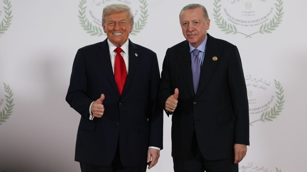 US President Donald Trump and Turkish President Recep Tayyip Erdogan stand side by side in dark suits, both giving a thumbs-up gesture against a white backdrop with official summit seals, framing the complex bilateral defense relations surrounding the F-35.