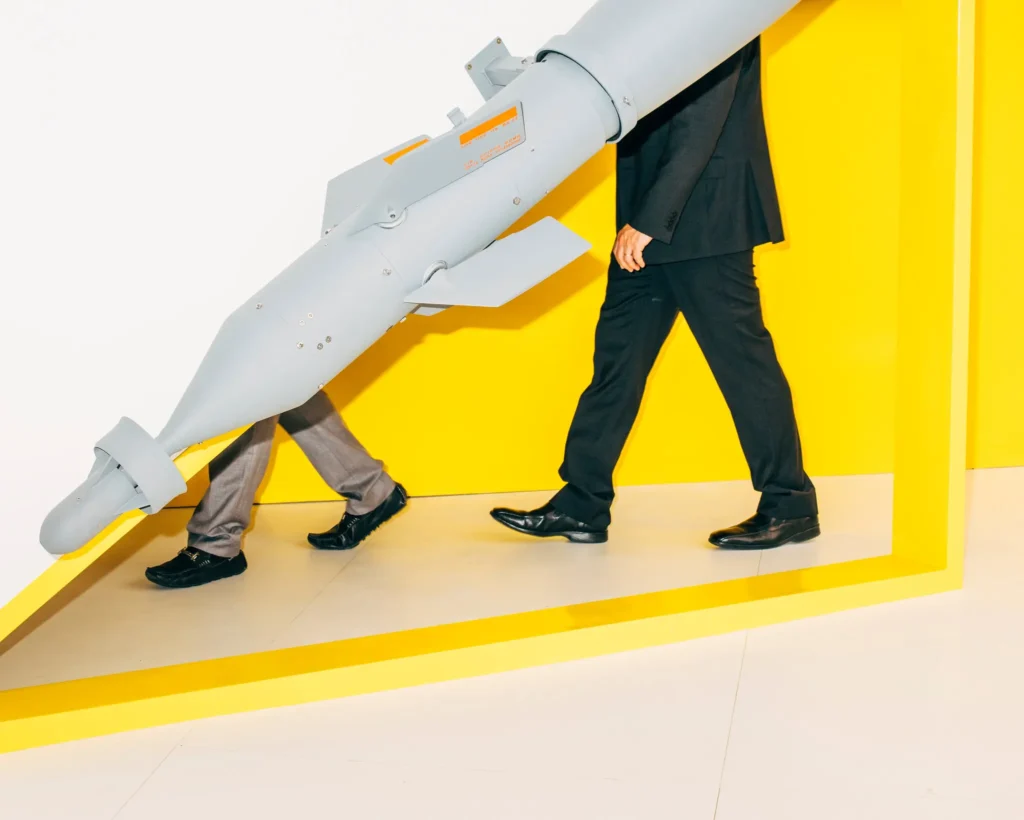 محاسبه استراتژیک نادرست: چگونه سه رهبر در سه جنگ گرفتار شدهاند Two men in business suits carrying a large gray guided missile through a bright white room framed by bold yellow geometric structures, symbolizing military escalation and Strategic Miscalculation.