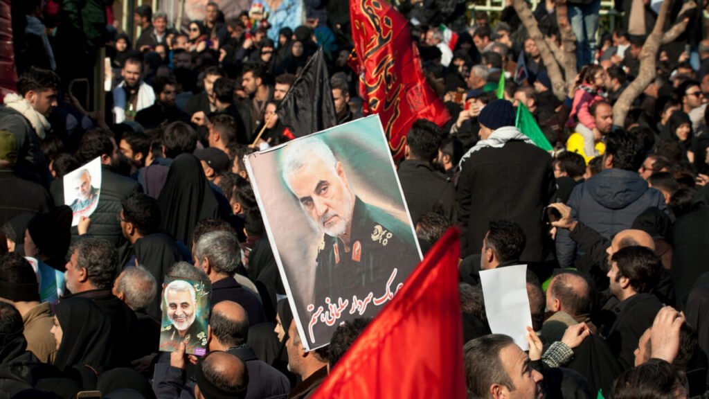 A large crowd of people packed together on a street carrying red and black flags and holding up posters of military commander Qasem Soleimani, providing visual context for geopolitical arguments regarding Iran Sanctions.