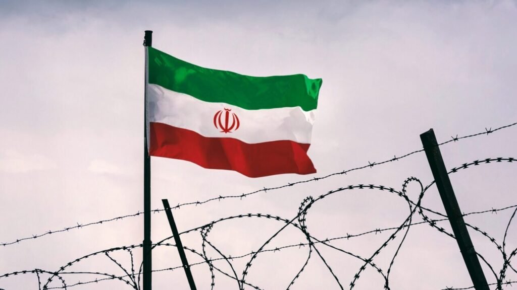 Can War Beat Sanctions in Limiting Iranian Nuclear Arms? The national flag of Iran waving in the wind behind a line of coiled barbed wire, symbolizing the geopolitical isolation and security tensions surrounding the development of Iranian nuclear arms.