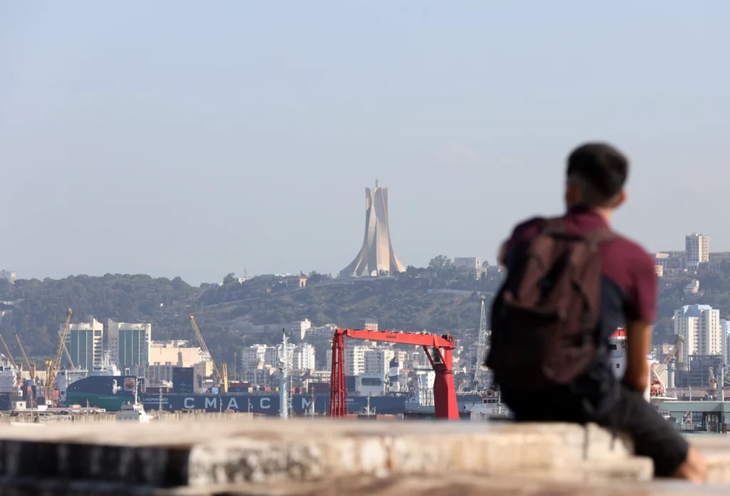 إصلاحات الجزائر المالية تُظهر تقدمًا. يجب على واشنطن أن تأخذ ذلك بعين الاعتبار A person with a backpack sits out of focus in the foreground overlooking the Port of Algiers with cargo ships and red cranes, while the Martyrs' Memorial stands prominently on the distant hillside, symbolizing economic development tied to Algeria's financial reforms.