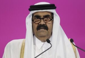 A close-up portrait of the late Hamad bin Khalifa al-Thani wearing traditional white headgear and glasses, speaking into microphones against a vibrant magenta background, commemorating the public life of the Emir.