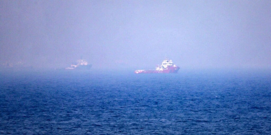 A wide, hazy shot of two commercial transport ships sailing on a vast blue ocean under a misty, overcast sky, highlighting the maritime trade routes affected by Trump's threats.