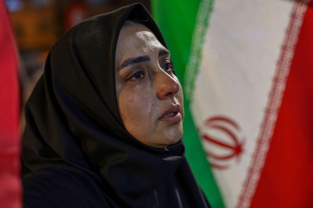 جنازة خامنئي ستختبر الدعم العام للجمهورية الإسلامية A close-up portrait of an Iranian woman wearing a black hijab with tears streaming down her face, looking upward with an emotional expression against a blurred national flag of Iran, capturing the public mourning context related to the Khamenei funeral.