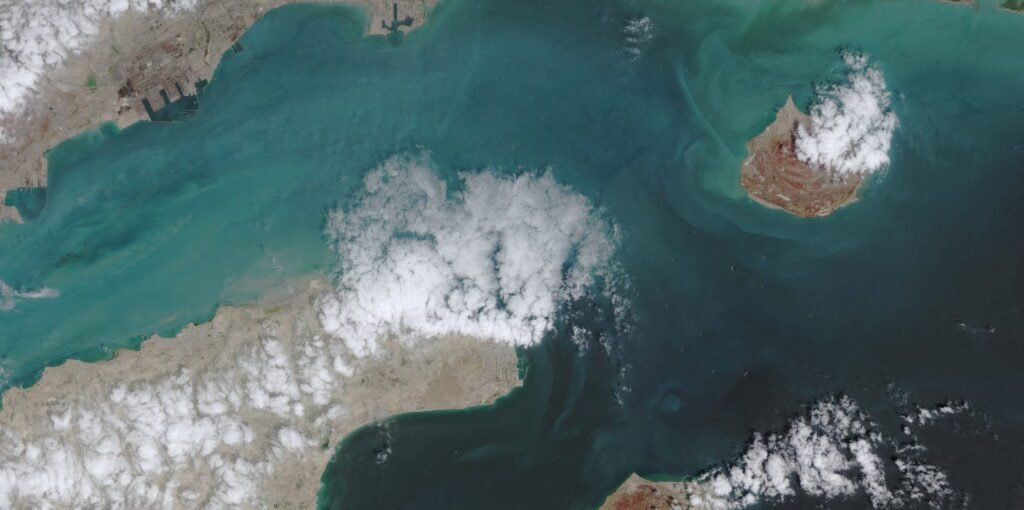 Will Hormuz be the American Suez? A high-altitude satellite view showing the Strait of Hormuz with coastal landmasses, distinct islands surrounded by varying shades of turquoise and deep blue seawater, and scattered white cloud formations above, providing geographic context for strategic discussions on whether it will become the American Suez.
