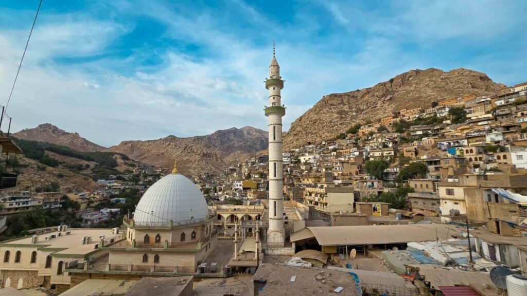درسهای کردستان برای جنگ ایران و نظم بینالمللی A high-angle scenic view over the historic mountain town of Akre featuring a white-domed mosque with a tall, slender minaret, surrounded by traditional stone houses built into the hillside under a blue sky, illustrating regional stability in Kurdistan.