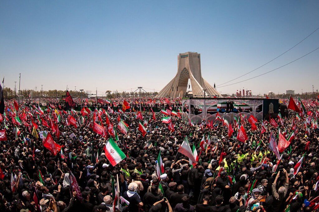 A wide-angle aerial shot capturing a massive crowd gathered in a large square in Tehran, featuring the Azadi Tower in the background and a sea of Iranian flags being held by attendees, reflecting the complex political atmosphere following the US-Israeli war against Iran.