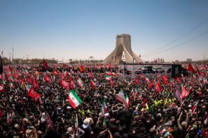 A wide-angle aerial shot capturing a massive crowd gathered in a large square in Tehran, featuring the Azadi Tower in the background and a sea of Iranian flags being held by attendees, reflecting the complex political atmosphere following the US-Israeli war against Iran.